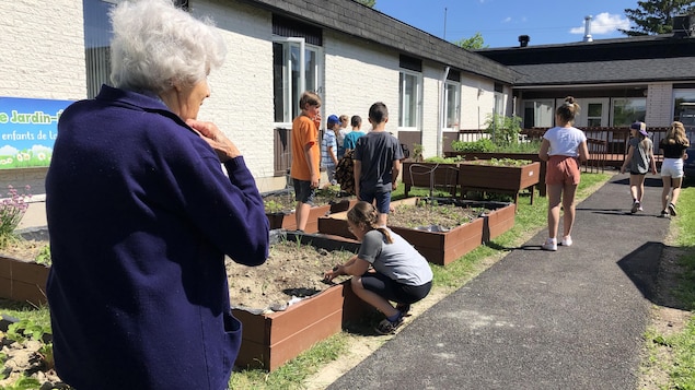 Une personne âgée regarde des jeunes dans un jardin.