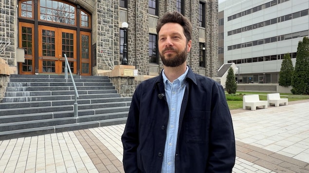 Un homme barbu portant une veste bleu marine sur une chemise en jean se tient debout à l'extérieur. Derrière lui, un grand bâtiment en pierre avec des marches d'entrée en granite et une porte en bois est visible, à côté d'un immeuble de bureaux moderne plus à l'arrière-plan. Le sol est dallé.