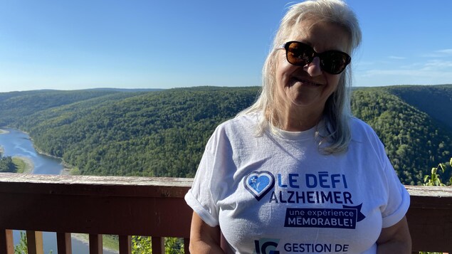 Doris Deschênes se trouve au belvédère de Saint-Alexis, on peut voir des montagnes et une rivière derrière elle.