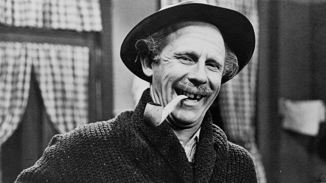 Le père Gédéon Plouffe (Doris Lussier), souriant, une pipe à la bouche.