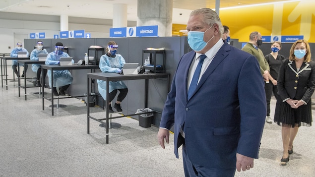 Doug Ford et la ministre de la Santé Christine Elliott devant des techniciennes qui font passer des tests à l'aéroport.