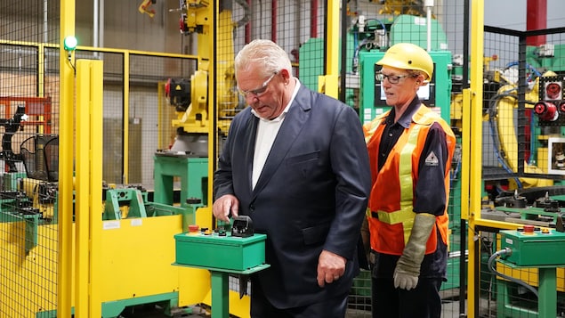 رئيس حكومة أونتاريو دوغ فورد يتفحص قطعة من المعدات في مصنع وهو يصغي إلى شرح من موظفة.