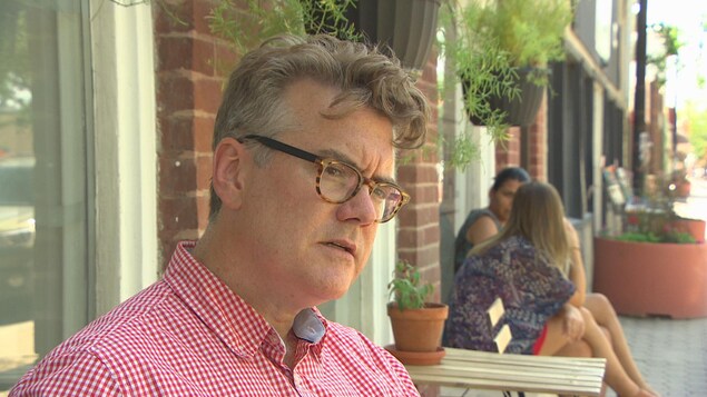 Un homme portant des lunettes et une chemise carreautée rouge et blanche est assis à la terrasse d'un café. Un homme portant des lunettes et une chemise carreautée rouge et blanche est assis à la terrasse d'un café.