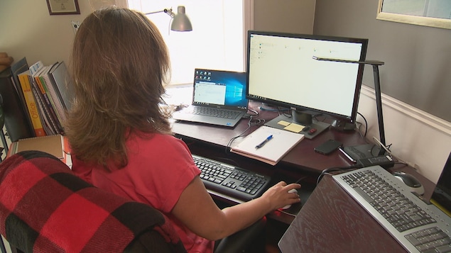 Une femme de dos est assise à un bureau. elle manipule de la main droite une souris d'ordinateur et regarde en direction des deux écrans qui sont devant elle.