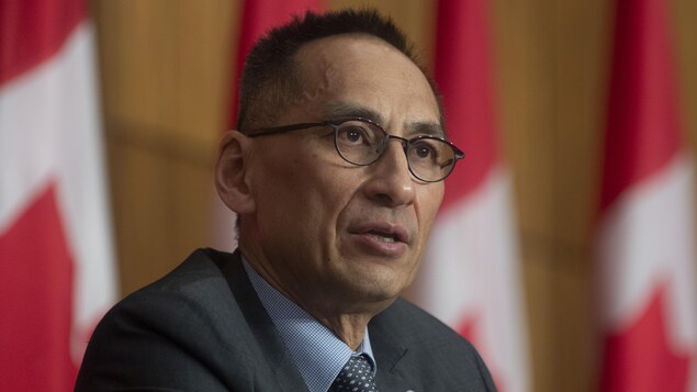 Howard Njoo nagsasalita sa isang press conference nasa likod niya ang mga watawat ng Canada.