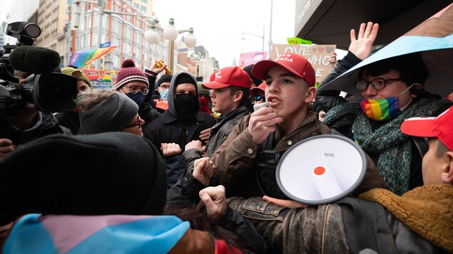 De jeunes hommes avec des porte-voix et des casquettes rouges « Save Canada » copiant le design des casquettes « MAGA »  sont entourés d'autres personnes avec des drapeaux arc-en-ciel.