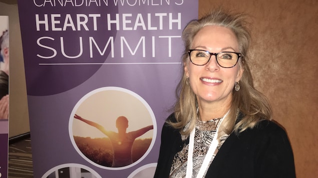 Une dame sourit à la caméra devant une bannière du Sommet canadien sur la santé cardiaque des femmes. Une dame sourit à la caméra devant une bannière du Sommet canadien sur la santé cardiaque des femmes.