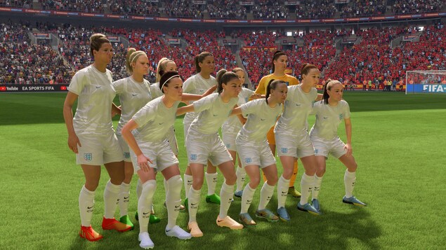 Dans un jeu, un groupe de femmes vêtues de maillots et de shorts de soccer posent en rangées, au milieu d'un terrain. Au loin, on voit des estrades remplies de personnes.
