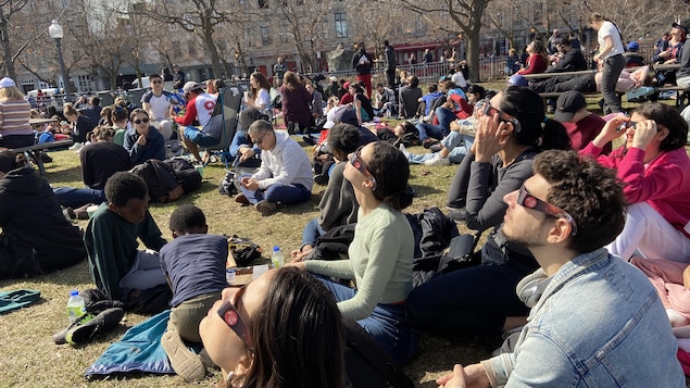 
أشخاص جالسون على العشب ويحملون نظارات كسوف الشمس وهم ينظرون إلى السماء.