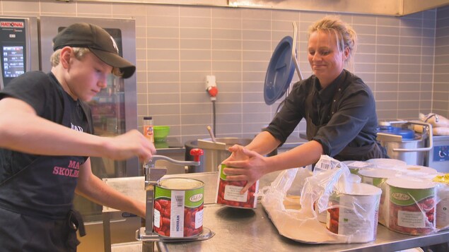 On voit Anne Marie et l'élève qui ouvrent de grosses boîtes de conserve dans la cuisine On voit Anne Marie et l'élève qui ouvrent de grosses boîtes de conserve dans la cuisine