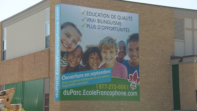 Un mur d'école en briques avec une grande affiche qui montre des jeunes souriants. Il est écrit que l'école ouvre en septembre.