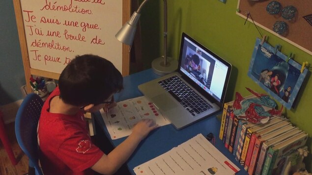 Un enfant écrit sur une feuille placée devant un écran d'ordinateur sur lequel on voit un autre enfant faire la même chose.