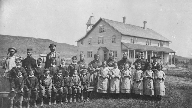 Photo d'archive où l'on voit des élèves et leurs superviseurs regroupés sur le terrain devant l'édifice. Des élèves garçons à gauche, des élèves filles à droite.