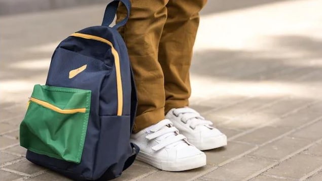 Un sac à dos bleu et vert se retrouve au pied d'une personne qui porte des souliers blancs et un pantalon beige.