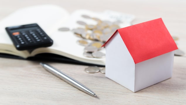 Une maison miniature repose près d'une calculatrice, un stylo et des pièces de monnaie.
