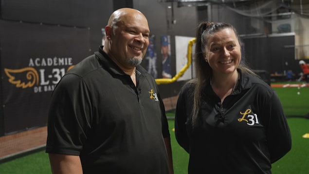 Un homme et une femme sourient dans un complexe où s'entraînent de jeunes joueurs de baseball. 
