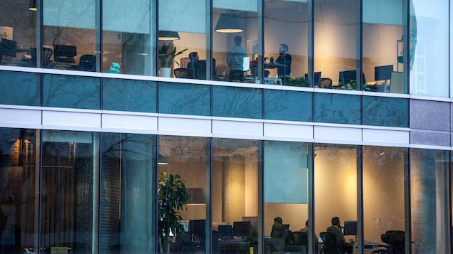 À travers la façade vitrée d'un bâtiment où se trouvent des bureaux, des gens travaillent.