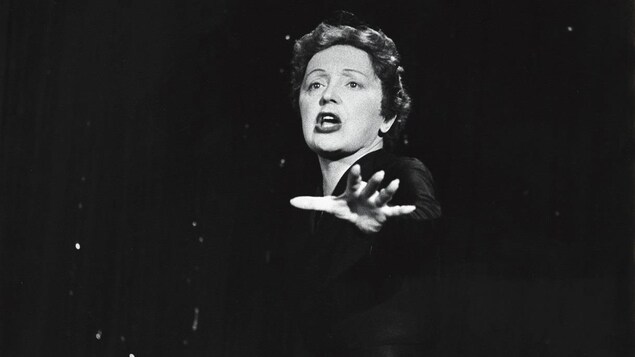 La chanteuse Édith Piaf, en performance, une main tendue devant elle.