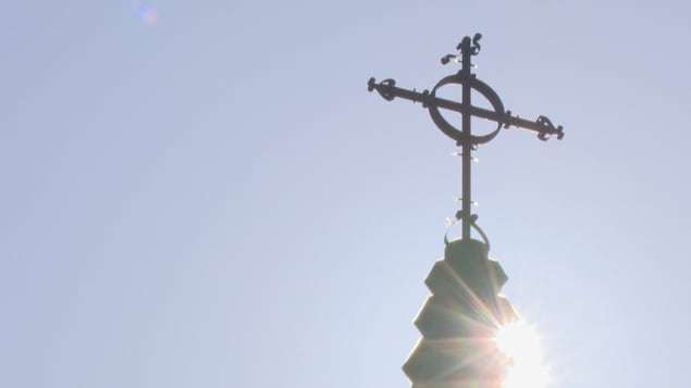 Une photo contre jour d'une croix au sommet d'une église. Le soleil brille derrière. Le ciel est bleu.