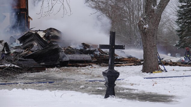 Une croix en métal se dresse dans la neige devant les cendres fumantes d'une église qui a brûlé.