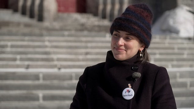 Une femme vêtue d'un manteau et d'une tuque sourit devant un parvis d'église. 