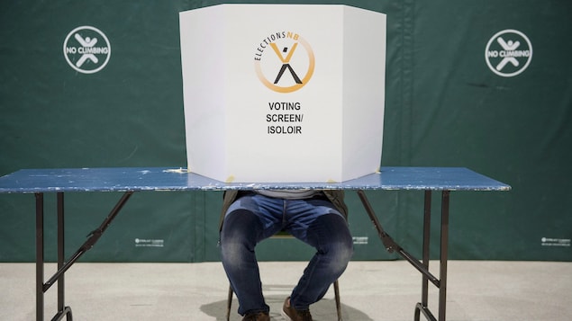 Un carton identifié par le logo d'Élections Nouveau-Brunswick cache le visage d'un électeur assis, dont on ne voit que les jambes.