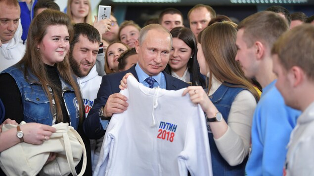 Une jeune femme offre un chandail au président Poutine. Ils sont entourés d'une vingtaine de jeunes qui sourient. Une jeune femme offre un chandail au président Poutine. Ils sont entourés d'une vingtaine de jeunes qui sourient.