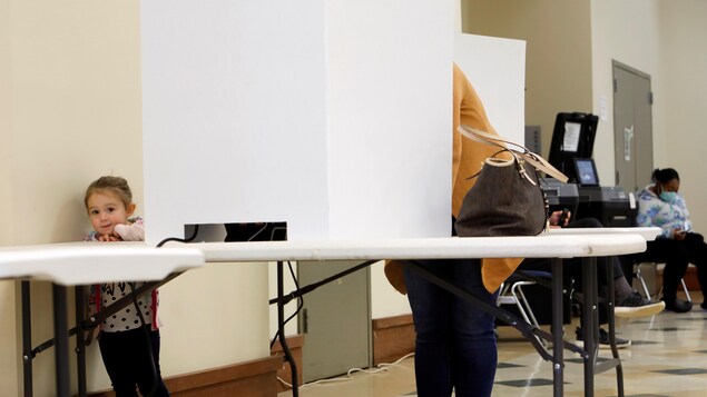 Une fillette regarde le photographe pendant que sa mère, cachée derrière le panneau blanc d'un isoloir, vote aux élections.