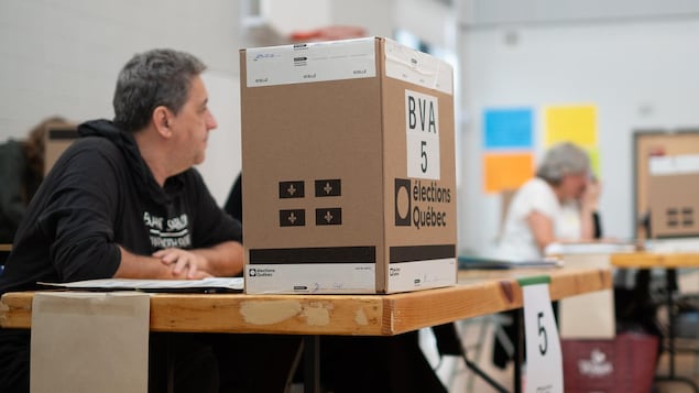 Une boîte de scrutin d'Élections Québec est posée sur une table, à côté d'un homme assis dans un bureau de vote.