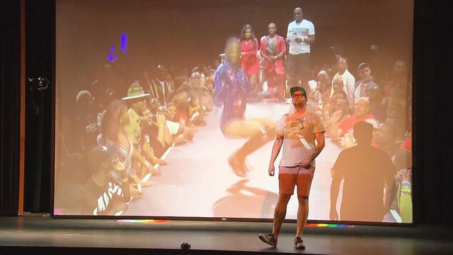 Une personne est debout sur scène dans un amphithéâtre sans spectateurs, lors d'une séance de répétition. On voit derrière un écran géant d'un enregistrement d'une personne qui danse.