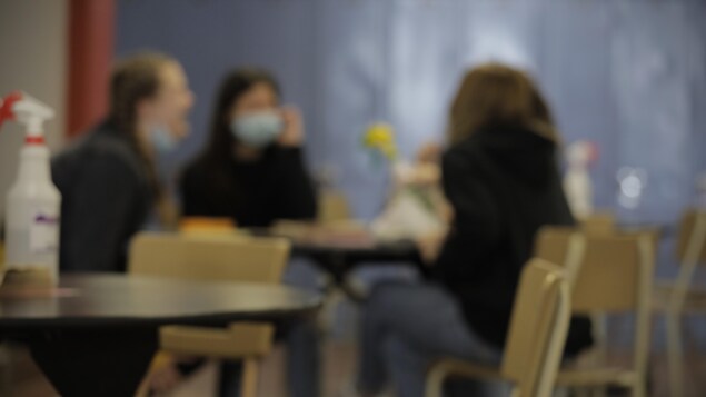 Des élèves sont assis à une table. Une est masquée, l'autre a le masque au dessous de la bouche.