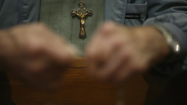 Un homme tient un chapelet entre ses mains durant la prière, à l'Église. Une croix pend à son cou.
