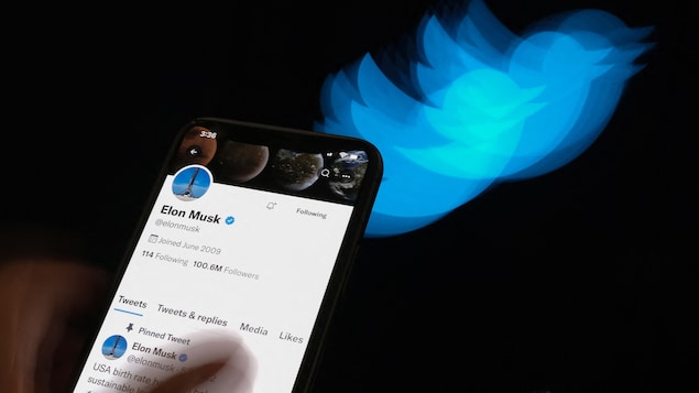 Un téléphone intelligent affiche la page d'accueil du compte Twitter d'Elon Musk, devant un écran noir sur lequel apparaît le logo du réseau social. 