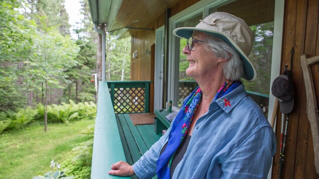 Une femme âgée debout sur la galerie de son chalet regarde vers la forêt.