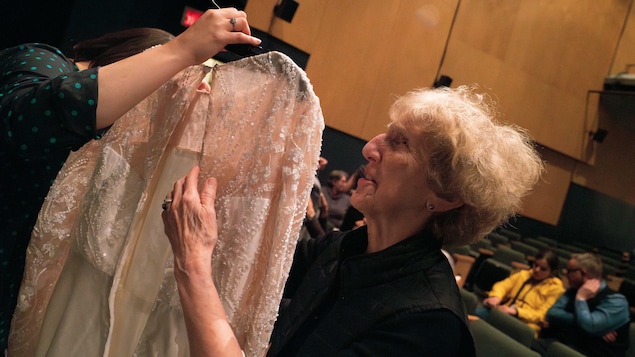 Une femme non voyante touche une robe de mariée sur un cintre qui est tenue par une femme dans une salle de spectacle.