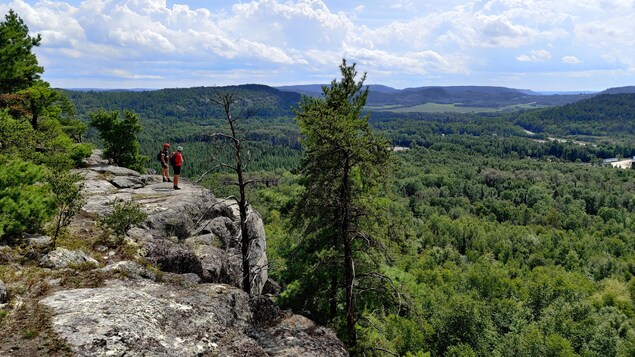 Deux hommes sont debout sur un cap de roche. Ils regardent les arbres à l'horizon et portent un casque de vélo.