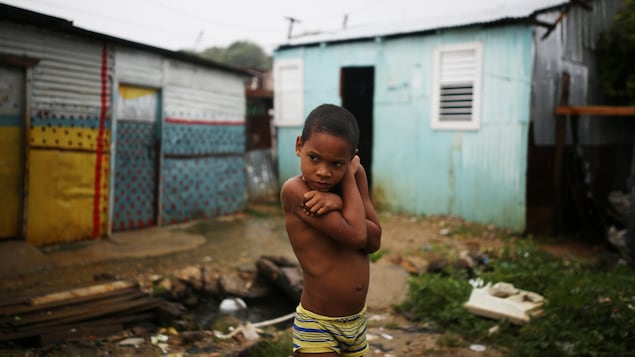 Un garçon se tient devant sa maison endommagée par l'ouragan, dans le quartier de La Cienaga, à Saint-Domingue, en République dominicaine. Il est vêtu d'un caleçon jaune et bleu, les bras enlacés autour de ses épaules.