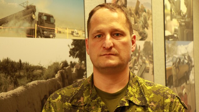 Un homme à l'air grave regarde la caméra devant une mosaïque de photos de l'armée canadienne. Un homme à l'air grave regarde la caméra devant une mosaïque de photos de l'armée canadienne.