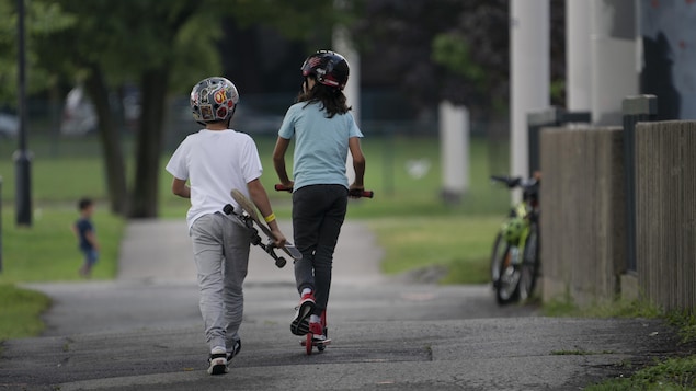Une fillette se déplace en trottinette, tandis qu'un garçon la suit en tenant une planche à roulettes.