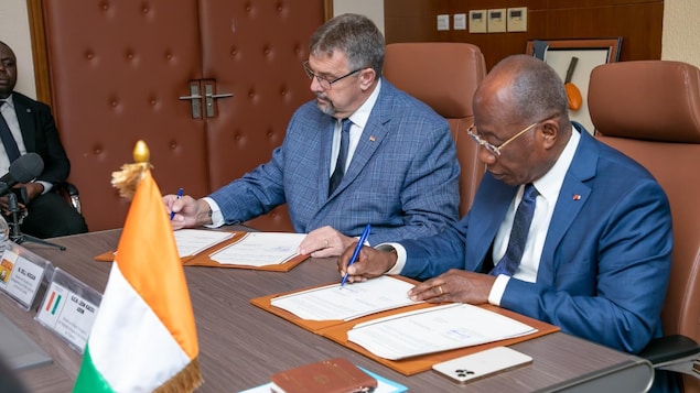 Assis à une table décorée d'un petit drapeau de la Côte d'Ivoire, deux hommes en complet signent des documents.