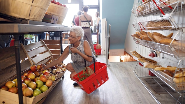 Une femme qui tient un petit panier d'épicerie s'accroupit pour prendre une pomme dans une caisse posée sur la rangée du bas, dans une épicerie de quartier.

