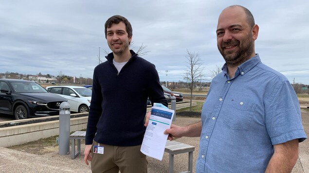 Deux hommes souriant dans l’entrée d’un stationnement tiennent en main un document.