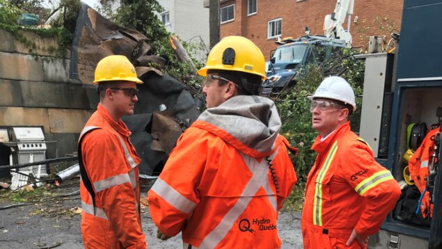 Trois hommes en combinaison orange discutent devant des débris.