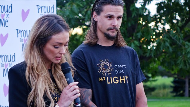 Une femme parle un micro pendant qu'un homme regarde le public devant une banderole où il est écrit « Proud to be me ». Une femme parle un micro pendant qu'un homme regarde le public devant une banderole où il est écrit « Proud to be me ».
