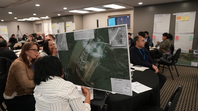 Image d'une salle de conférence avec au premier plan une participante tenant une grande carte avec une vision satellite d'une communauté.