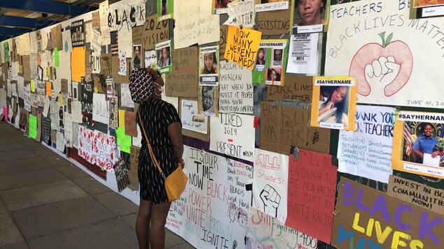Une jeune femme observe des affiches collées sur un mur.