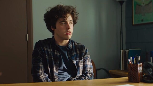 Un jeune homme avec une chemise carreautée assis à un bureau.