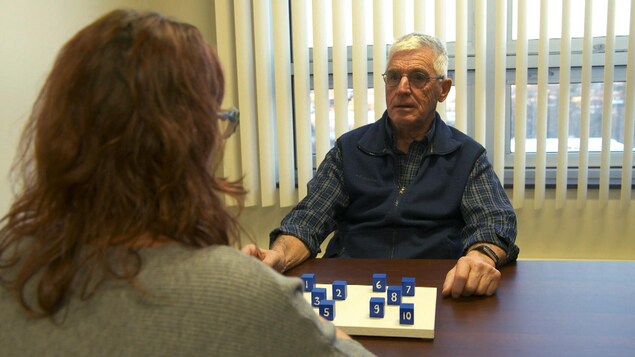 On voit un homme de face assis à une table devant une femme qui lui administre le texte. Devant eux sont posés des cubes bleus numérotés.
