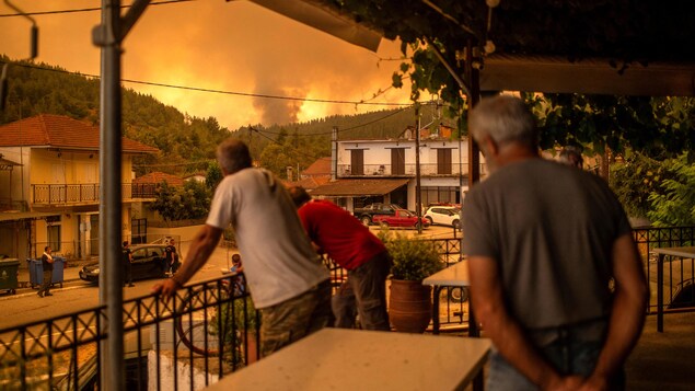 Trois hommes sont accoudés à la rambarde d'une terrasse du village et regardent vers les fumées qui s'échappent de l'incendie de l'autre côté de la colline.