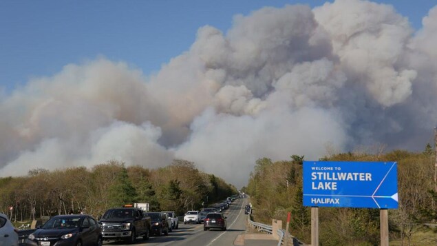Une file de voitures en rang sur l'autoroute près de l'affiche qui souhaite la bienvenue à Stillwater Lake. Une épaisse fumée de feu de forêt couvre le ciel.